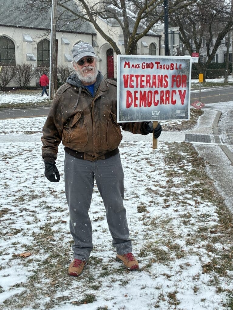 Man holding 'Veterans for Democracy' sign in snow.