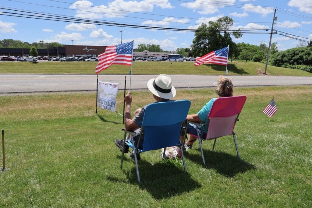 Two people sitting on lawn chairs waving American flags roadside