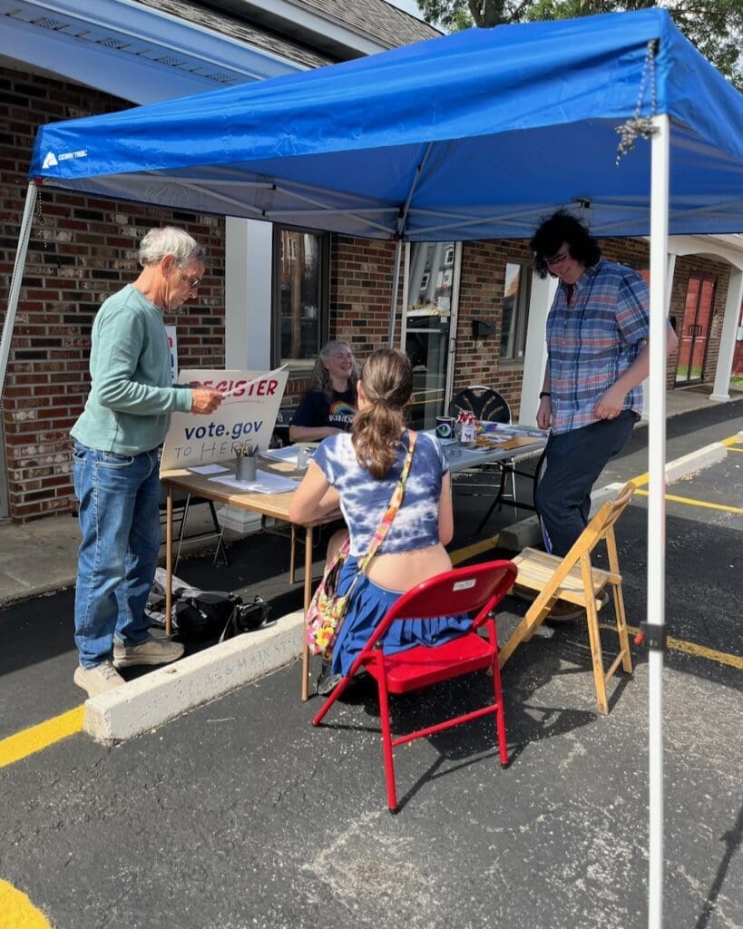 Volunteers at outdoor voter registration table under blue canopy