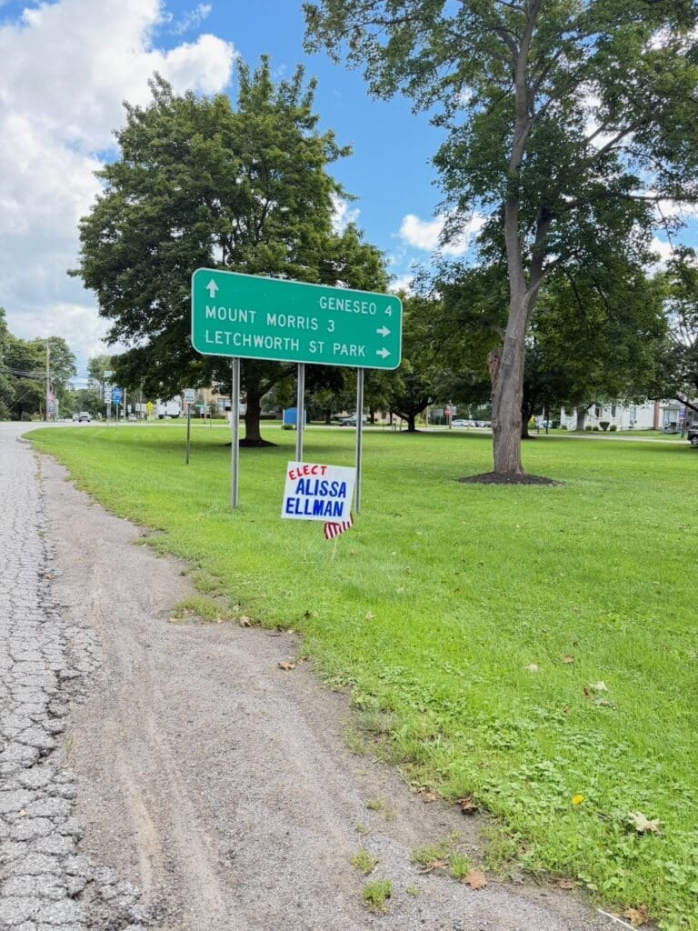 Green road sign for Geneseo and Letchworth State Park with campaign sign